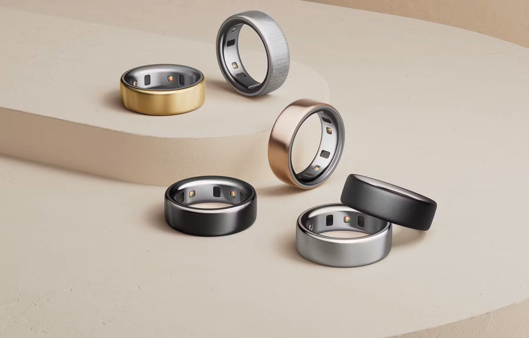Six Oura rings in gold, silver, black, and rose gold on a pale pedestal